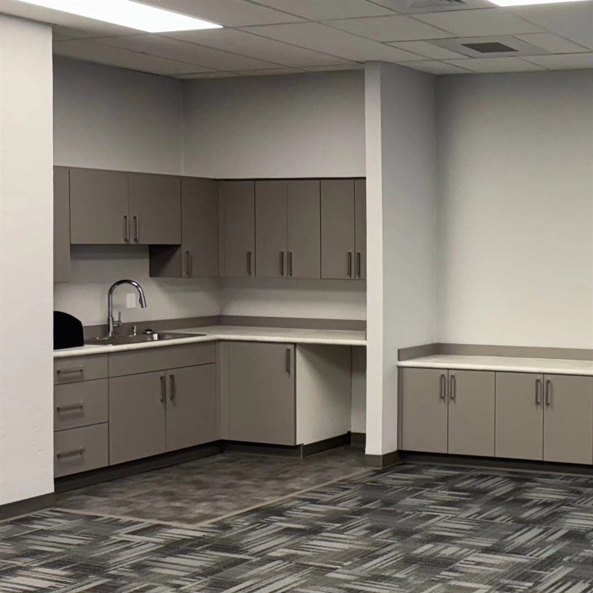 Office kitchenette with taupe slab-front casework, upper cabinets and a corner desk return over carpet tile