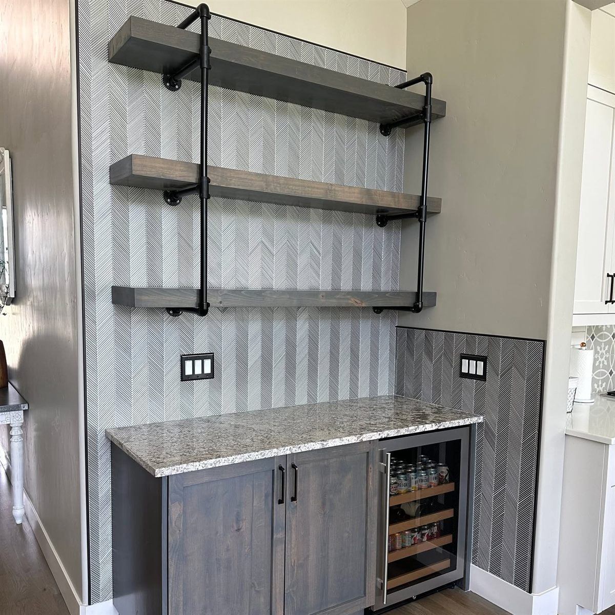Custom dry bar with industrial bracket floating shelves above a granite counter, a built-in wine fridge and a herringbone tile backsplash